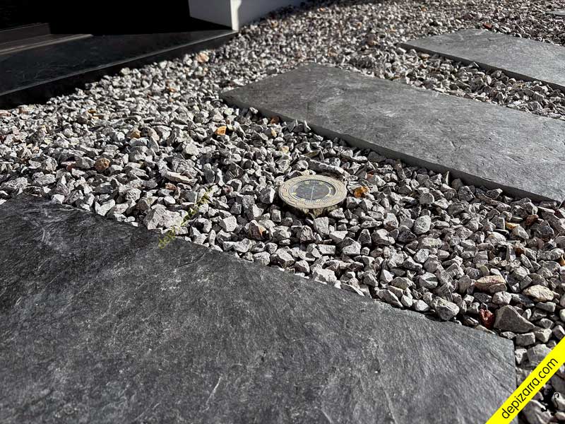 **Nombre de archivo:** ``` losas-pizarra-grava-luz-solar-jardin.jpg ``` **Título:** ``` Losas de pizarra y grava con luz solar decorativa en jardín ``` **ALT:** ``` Losas de pizarra sobre grava con luz solar decorativa en jardín exterior