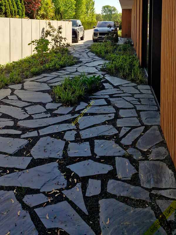 Pavimento de pizarra natural irregular en el acceso a una vivienda, con vegetación entre las piezas.