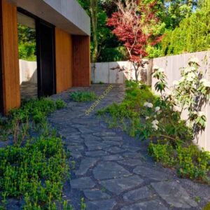 Pavimento de pizarra natural irregular en un jardín, rodeado de vegetación junto a una vivienda moderna.
