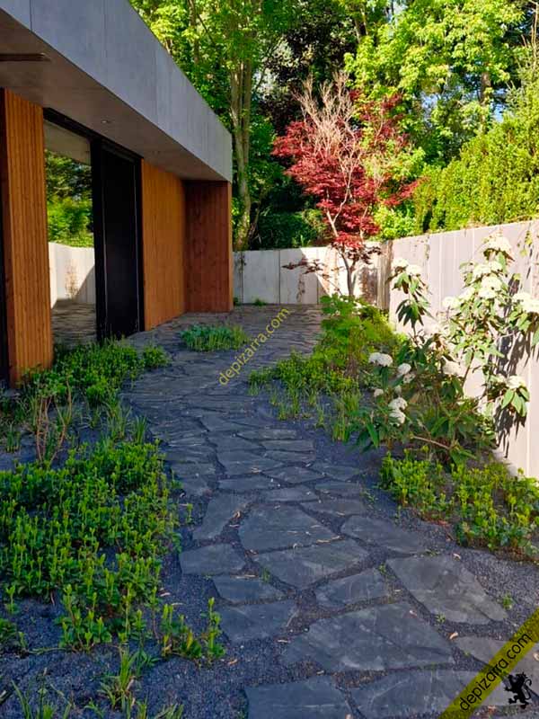 Pavimento de pizarra natural irregular en un jardín, rodeado de vegetación junto a una vivienda moderna.